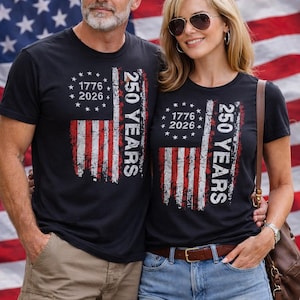 Puede incluir: Camisetas negras con un dise&ntilde;o desgastado de la bandera estadounidense. Las camisetas presentan el texto "1776 2026" y "250 YEARS" en blanco. El fondo es una bandera estadounidense.
