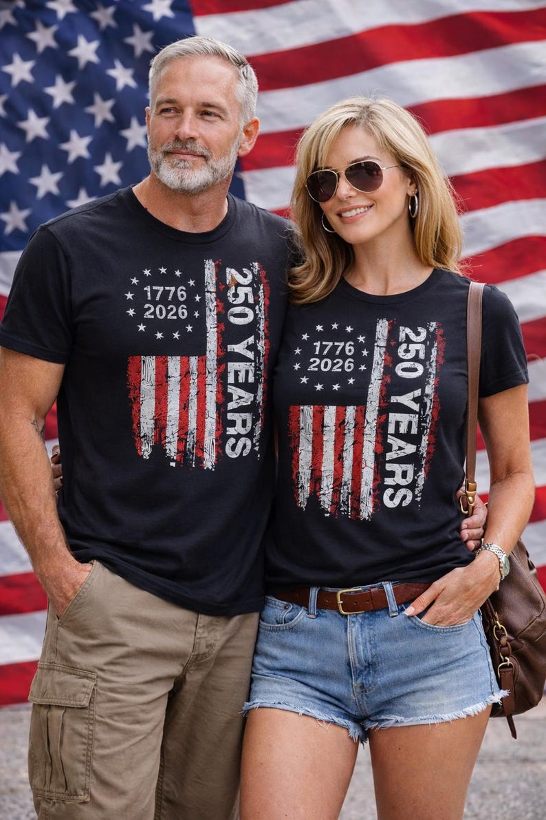 Puede incluir: Camisetas negras con un dise&ntilde;o desgastado de la bandera estadounidense. Las camisetas presentan el texto "1776 2026" y "250 YEARS" en blanco. El fondo es una bandera estadounidense.