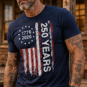 Puede incluir: Camiseta azul marino con un dise&ntilde;o desgastado de la bandera estadounidense. El texto dice "1776 2026" y "250 YEARS" en blanco y rojo. La camiseta tiene mangas cortas y cuello redondo.