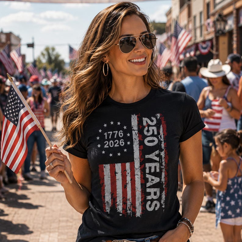 Puede incluir: Camiseta negra con un dise&ntilde;o desgastado de la bandera estadounidense. El gr&aacute;fico de la bandera incluye el texto "1776 2026" y "250 YEARS". La persona de la imagen sostiene una peque&ntilde;a bandera estadounidense.