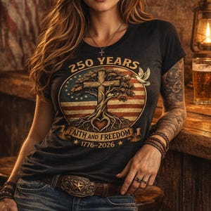 May include: A dark gray t-shirt with a graphic celebrating 250 years of faith and freedom. The design includes a cross, tree, and American flag elements. The person is wearing a brown cowboy hat and belt.