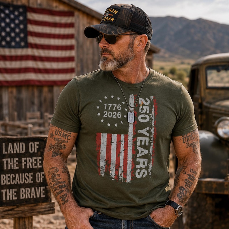 Puede incluir: Camiseta verde oliva con un dise&ntilde;o de bandera estadounidense desgastada y el texto "250 YEARS 1776-2026". La camiseta tambi&eacute;n presenta el texto "USMC". La persona lleva una gorra negra con "VIETNAM VETERAN".