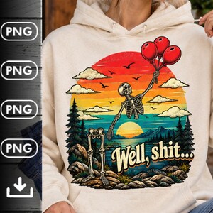May include: A cream-colored hooded sweatshirt featuring a graphic print of a skeleton floating with red balloons over a landscape with a lake, mountains, and pine trees. The text "Well, shit..." is printed in a bold, stylized font below the skeleton.