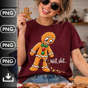 May include: A maroon short-sleeve t-shirt featuring a graphic of a sad gingerbread man with a missing leg and the text "Well, shit..." printed in white. The gingerbread man has a green bowtie, red and green buttons, and white icing trim.