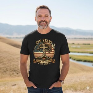 May include: Black t-shirt with a circular graphic featuring a tree with a cross, the American flag, and the text "250 Years Faith and Freedom 1776-2026". A dove is on the right side of the graphic. The person is wearing a watch.