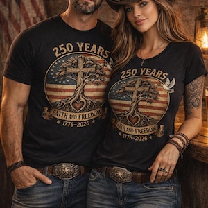 May include: Two people wearing matching black t-shirts with a patriotic design. The shirts feature a cross, tree, and American flag graphic with the text "250 YEARS FAITH AND FREEDOM 1776-2026". They are wearing cowboy hats, jeans, and belts.
