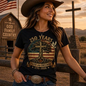 May include: A black t-shirt with a circular graphic featuring a cross, tree, and American flag design. The text reads "250 YEARS" and "FAITH AND FREEDOM 1776-2026". The person is wearing a brown cowboy hat and belt.