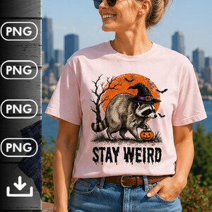 May include: A light pink t-shirt featuring a graphic of a raccoon wearing a black witch hat and carrying a small orange jack-o'-lantern. The background shows a large orange moon, bare trees, and bats. The text STAY WEIRD is printed in black.