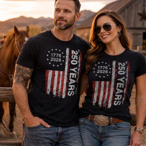 Puede incluir: Camisetas negras con un dise&ntilde;o desgastado de la bandera estadounidense. Las camisetas presentan el texto "1776 2026" y "250 YEARS" en blanco. Las personas visten vaqueros azules y est&aacute;n de pie frente a una valla de madera.