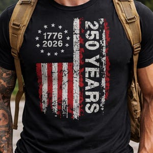 Puede incluir: Camiseta negra con un dise&ntilde;o desgastado de la bandera estadounidense con rayas rojas y blancas y estrellas. La camiseta incluye el texto "1776 2026" y "250 YEARS" en blanco, celebrando un hito hist&oacute;rico.