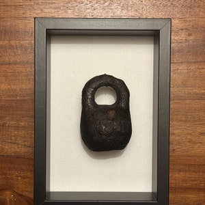 May include: A dark brown, antique-looking, metal kettlebell with a rounded shape and a small handle on top, displayed in a black frame on a white background.
