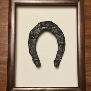 May include: A framed horseshoe with a dark brown frame. The horseshoe is made of metal and has a rustic appearance.