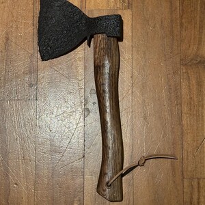 Può includere: Un'ascia nera con manico in legno e un cinturino in pelle. L'ascia ha una lama leggermente curva e una testa arrotondata.