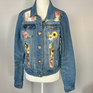 Può includere: Giacca di jeans blu chiaro con ricami floreali. La giacca presenta fiori ricamati in rosa, giallo e bianco, con pannelli trasparenti. Ha un classico design con bottoni e bottoni color rame.