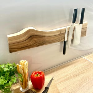 May include: A wooden knife holder with three knives and a white paper towel roll. The holder is made of light brown wood with a wavy design and a white light strip. The knives have black handles and silver blades.