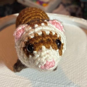 May include: A handmade crochet ferret toy. The ferret has a brown body and head markings, with white and pink accents on the face and ears. It has black safety eyes and a pink nose. The ferret is made of yarn.