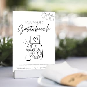 May include: A white card with the words "POLAROID Gästebuch" in elegant script, featuring a line drawing of a camera with a heart. The card also includes the phrases "FOTO KNIPSEN, FOTO EINKLEBEN, WÜNSCHE HINTERLASSEN" and "Danke, dass du unseren Tag unvergesslich".