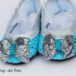 Può includere: Un paio di scarpine da bambino con un design scintillante. Le scarpe sono principalmente turchesi con accenti glitterati argentati sulla punta e sul tallone. Ogni scarpa ha un grande fiocco argentato con un nastro turchese. Su sfondo bianco.