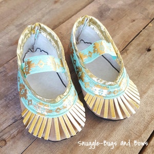 Può includere: Scarpine da bambino con un design oro e turchese. Le scarpe hanno una frangia dorata intorno alle dita e un cinturino turchese con ricami dorati. Le scarpe sono su una superficie di legno. Il testo "Snuggle-Bugs and Bows" è visibile.