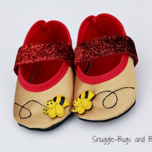 Puede incluir: Un par de zapatos de bebé con temática de abeja. Los zapatos son de color beige con ribete rojo y una correa roja con purpurina. Cada zapato tiene una aplicación de abeja amarilla y negra y un diseño de línea negra cosida. El texto "Snuggle-Bugs and Bows" está en la parte inferior.