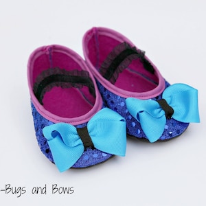 Può includere: Un paio di scarpine da bambino con esterno blu con paillettes e interno rosa acceso. Ogni scarpa ha un grande fiocco azzurro e un cinturino nero sulla parte superiore. Il testo "Snuggle-Bugs and Bows" è in basso.