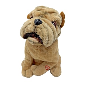 May include: A plush, tan-colored Shar Pei dog toy with a wrinkled face, floppy ears, and a black nose. The toy has a red button on its paw and is sitting upright.