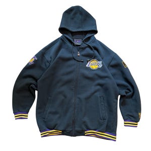 May include: A navy blue zip-up hoodie with a hood and the Los Angeles Lakers logo on the chest and sleeve. The jacket has purple and yellow striped trim on the cuffs and hem. The jacket has a zipper closure.