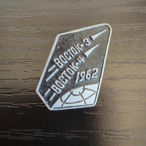 May include: A silver-toned metal pin with a black background featuring the text "BOSTOK-3" and "BOSTOK-4" stacked vertically, the year "1962" to the right, and a stylized graphic of a globe with grid lines at the bottom of the diamond-shaped badge.