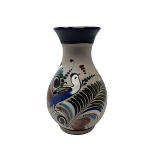 Puede incluir: Un jarrón de cerámica con cuerpo gris y borde azul oscuro. El jarrón presenta un diseño pintado a mano de un pájaro con plumas azules, blancas y marrones, rodeado de hojas estilizadas y elementos florales. El jarrón mide aproximadamente 20 cm de alto.