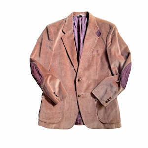 Op de afbeelding: Een roze corduroy blazer met een gekerfde revers, twee voorzakken en knopen. De ellebogen hebben donkerpaarse patches. De binnenvoering is dieppaars.
