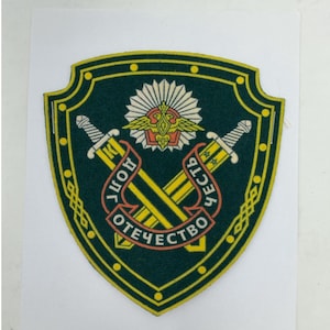 May include: A shield-shaped embroidered patch in dark green with a yellow border. It features two crossed swords, a central golden eagle emblem, and a white sunburst. A red ribbon banner displays the Cyrillic text DOLL, CHEST, OTECHESTVO.