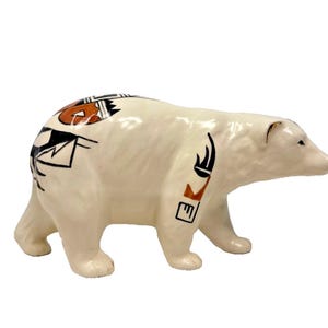 May include: A ceramic polar bear figurine with a glossy off-white finish. The bear features hand-painted geometric patterns in black and burnt orange on its back and front leg, with a smooth, sculpted body and a forward-facing head position.