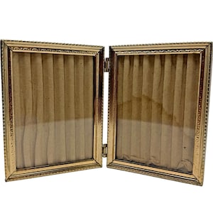 Peut inclure: Un cadre photo double doré à charnière. Le cadre est rectangulaire avec une bordure décorative. L'intérieur de chaque cadre a un support beige. Le cadre est ouvert, révélant l'intérieur. Le cadre est probablement en métal.