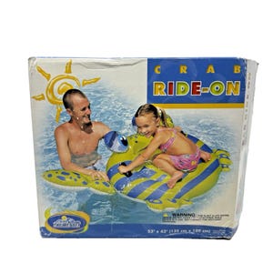 Pode incluir: Caixa para um flutuador insuflável em forma de caranguejo. O flutuador é amarelo e azul, com as palavras "CRAB RIDE-ON" em azul e vermelho. A caixa mostra um homem e uma menina numa piscina, a andar no flutuador. O flutuador mede 135 cm x 109 cm.