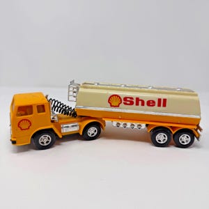 May include: A vintage toy Shell tanker truck. The cab is orange with the Shell logo, and the trailer is cream and orange with the Shell logo and text. The truck has black wheels and a silver ladder.
