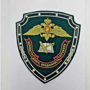 May include: A dark green shield-shaped patch featuring a gold double-headed eagle emblem at the top. Below, an open book with a sword and wheat stalk. A red ribbon banner displays the Cyrillic text: ВЕРНОСТЬ ДОЛГУ, ПРЕДАННОСТЬ ОТЧИЗНЕ.