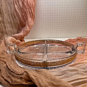 May include: A clear glass serving tray with four compartments and two handles. The tray has a gold-colored band with an ornate design around the rim and a blue accent line. The tray is on a brown and pink fabric.