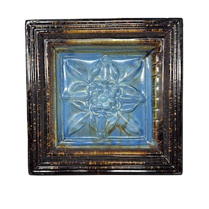 May include: Square ceramic tile with a raised floral design in shades of blue. The tile is framed with a dark brown border with a textured appearance. The center features a detailed flower with rounded petals and a central cluster.