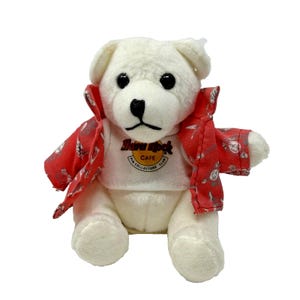 Puede incluir: Un oso de peluche blanco con una chaqueta roja estampada y una camiseta blanca con el logo de Hard Rock Cafe. El oso tiene ojos negros y una nariz negra. Un artículo de colección.