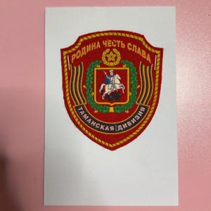 May include: A shield-shaped embroidered patch featuring a red background with gold borders. It displays a central emblem of a rider on a white horse, surrounded by a green wreath and a gold star. Cyrillic text reads RODINA CHEST SLAVA TAMANSKAYA DIVIZIYA.