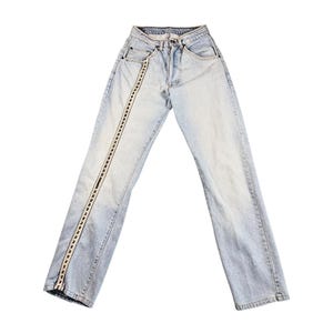 May include: Light-wash blue denim jeans featuring a high-waisted fit, a button fly closure, and a decorative vertical strip of white and black woven trim running down the length of the right pant leg from the waistband to the hem.