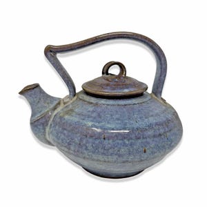 Puede incluir: Tetera de cerámica hecha a mano con un esmalte azul y morado. La tetera presenta un asa curvada, un pico vertedor y una tapa con un pequeño lazo. La forma general es redondeada.