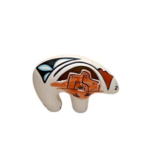 May include: A ceramic bear figurine with a cream-colored body. The bear features a black and white design on its back, with blue accents. An orange and brown pueblo scene is painted on the bear's side. The figurine has a small, rounded head and a stubby leg.