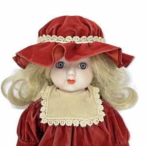 Può includere: Una bambola di porcellana vintage con capelli biondi e occhi blu. La bambola indossa un vestito di velluto rosso e un cappello abbinato con un bordo in pizzo color crema. Il viso della bambola è dipinto con guance rosee e labbra rosse.