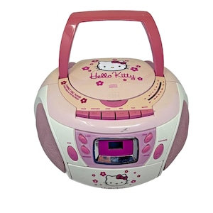 Puede incluir: Un radiocasete Hello Kitty rosa y blanco con asa. El reproductor de discos compactos tiene una pantalla digital y botones para varias funciones. El radiocasete presenta el logotipo de Hello Kitty y detalles florales.