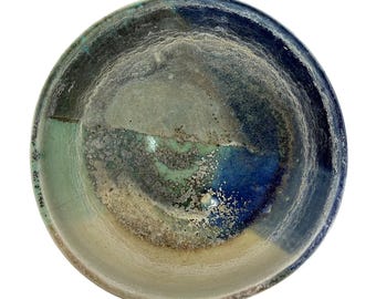 May include: A round ceramic bowl featuring a reactive glaze with blended shades of deep navy blue, seafoam green, and sandy beige. The interior surface displays a textured, mottled finish with crystalline patterns and a smooth, glossy rim.