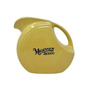 May include: A yellow ceramic pitcher with a rounded body and a handle. The pitcher has a spout for pouring and a black logo that reads "Millennium The Year 2000". The pitcher is a vintage collectible.