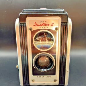 May include: A vintage Kodak Duaflex II camera featuring a black and silver rectangular body with vertical ribbed side panels. The front face displays the text Kodak Duaflex II Camera and Kodet Lens, with two circular glass lenses stacked vertically.