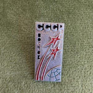 Puede incluir: Un pin rectangular de metal plateado con el texto CCCP en la parte superior y VOSTOK verticalmente a la izquierda. Dos estrellas rojas con los números 5 y 6 se muestran con estelas sobre un gráfico de un globo parcial abajo.
