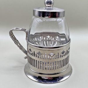 Op de afbeelding: Een zilveren suikerdispenser met een glazen pot en een decoratief handvat. De pot heeft een zilveren deksel met een bolvormige knop. De basis van de dispenser heeft een decoratief, opengewerkt ontwerp.